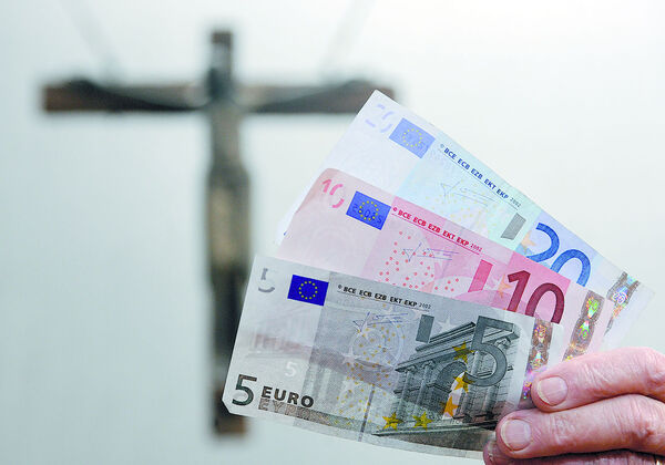 Eine Hand mit Euro-Geldscheinen vor einem Kreuz.