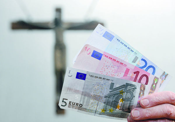 Eine Hand mit Euro-Geldscheinen vor einem Kreuz.