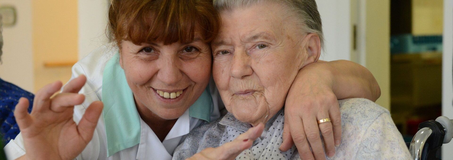 Pflegerin lächelt mit Seniorin in die Kamera