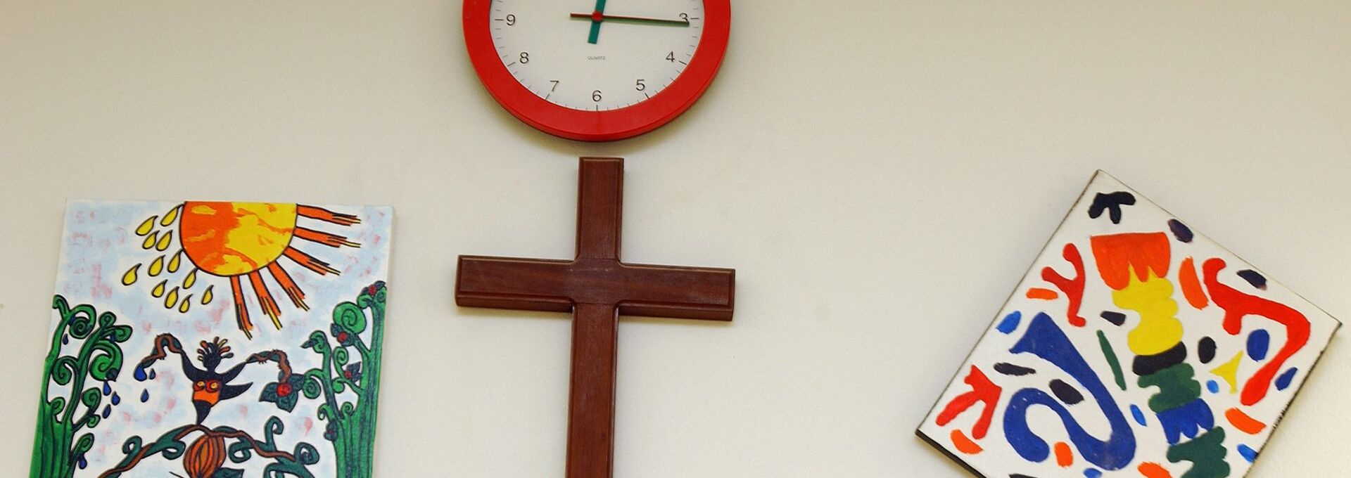 In dem Klassenzimmer einer Schule hängen über der Türe ein Kreuz, eine Uhr und zwei gemalte Bilder.